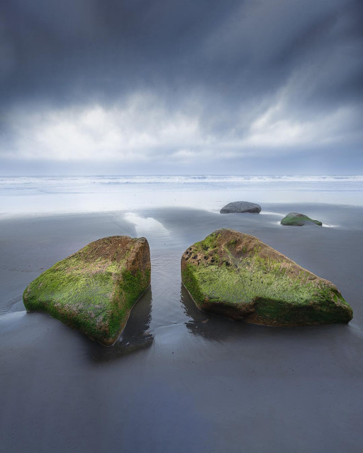 A vertical, fine-art landscape photograph of a moody, overcast black sand beach at Rapanui Beach, Taranaki. In the immediate foreground, two large, matching boulders covered in vibrant green moss sit close together, separated by a narrow channel of reflective, wet sand carved by the tide. Further back on the smooth, dark shore, another smaller pair of mossy rocks rests near the breaking white surf of the Tasman Sea. The sky above is filled with heavy, dark grey storm clouds, creating a somber, minimalist, and atmospheric tone.
