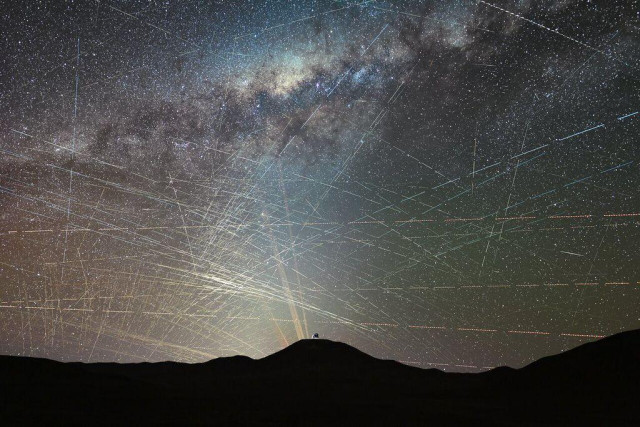 Time-lapse photo of the night sky over Paranal Observatory in Chile, with the observatory seen as a bump on the distant silhouette of the hills. Overhead, the glow of the Milky Way dominates the upper half of the sky while numerous satellite trails cover the sky.