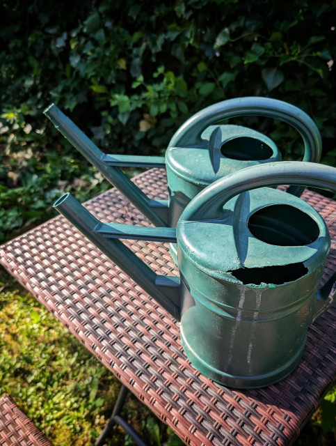 Zwei identische grüne Gießkannen auf einem Gartentisch, eine ganz, die andere mit Löchern Bei dieser wurde ein Stück der Tülle abgesägt und als Ersatzteil verwendet