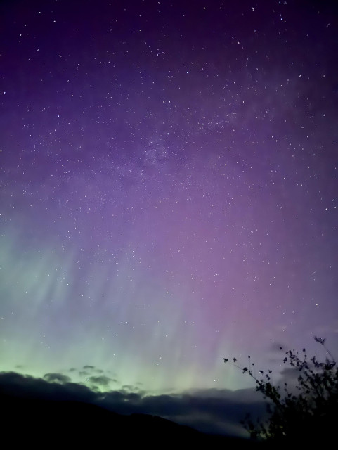 A night sky filled with stars, displaying shades of purple and green, with silhouettes of clouds and a dark landscape at the bottom.