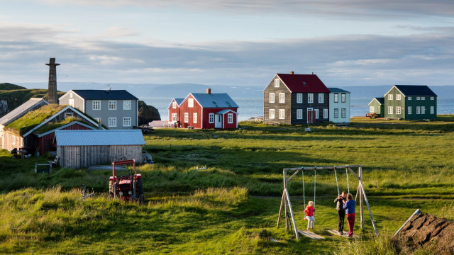 an image of a small village in Iceland, a few houses on green grass, a swingset with some kids. The blue sea is visible in the background
