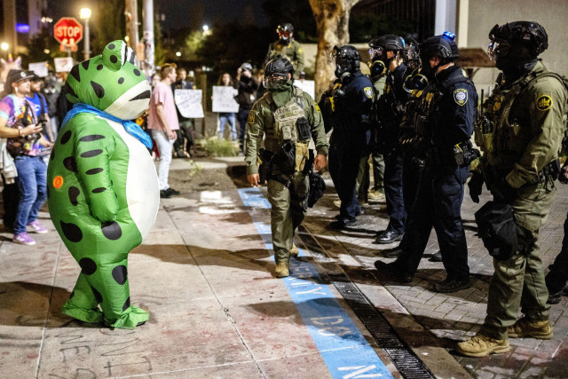 A person in a frog costume being confronted by armed police and soldiers at a No Kings rally.