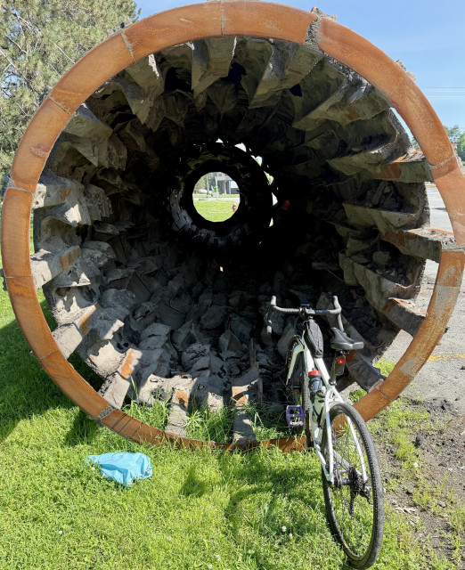An old part  of an asphalt or cement machine at a gravel pit. Son's old Salsa gravel bike is propped up near one end leaning on a large metal tube that appears to have inward angled teeth all along the walls.