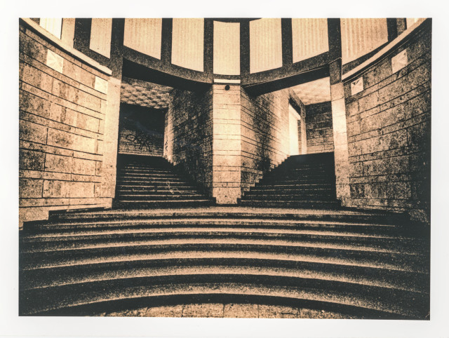 A moody lith print showing a symmetrical underground stairwell, where curved steps and towering stone walls frame two dark passages leading toward soft light. The coarse grain and warm sepia tones give the scene an eerie, timeless feel.