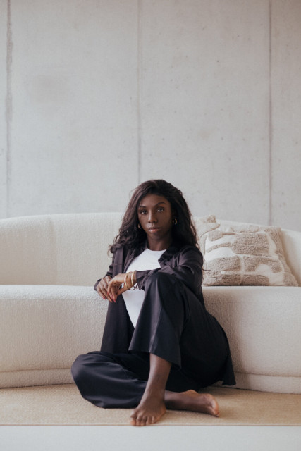 A Black woman with long wavy dark hair sits barefoot cross-legged on a light beige rug in front of a cream-colored textured sofa. She wears a white t-shirt beneath a loose-fitting black satin pantsuit with wide-leg trousers and matching blazer. Gold jewelry includes hoop earrings, stacked bangles on her right wrist, and rings. Her posture is relaxed yet poised with one arm resting on her bent knee, hands gently clasped. She gazes directly at the camera with a calm, confident expression. The setting features minimalist decor including a textured geometric pillow on the sofa behind her and a neutral concrete-textured wall in the background, creating a serene, modern atmosphere with warm natural lighting.