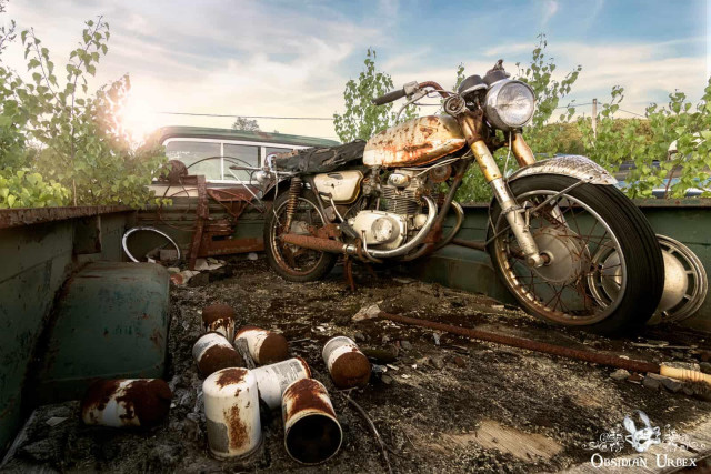 A rusted vintage motorcycle rests in the bed of an abandoned pick-up truck, surrounded by other decaying items under a bright sky.