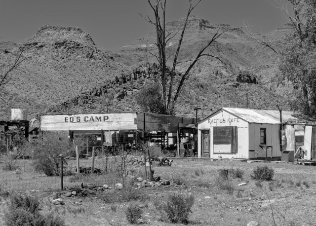 A black and white landscape photo of a long abandoned roadside stop with gas station and a cafe. The buildings are painted white. There's a small water tower on the left. The background are rugged desert mountains. The branches of a long dead tree are seen directly behind the buildings. The sky is clear.