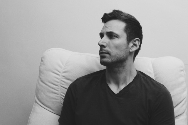 A man is sitting on a white chair, looking thoughtfully to the side. The image is in black and white, highlighting his facial features and expression. The background is plain and light-colored.