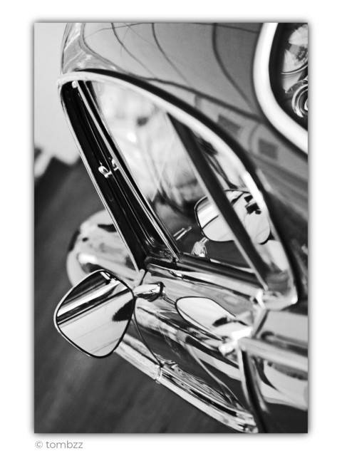 Black and white photograph showing a close-up detail of a classic car. The frame is filled with smooth, flowing body lines, the chrome trim around the side window, and a small chrome side mirror mounted on the fender. In the upper right corner, part of a round headlight is visible, its polished bezel contrasting with the darker bodywork. The reflections across the highly polished surface immediately draw the eye. Distorted outlines of nearby buildings and tree branches stretch across the car like an abstract drawing. The chrome reflects the light with almost mirror-like precision, creating a rich interplay of bright highlights and deep shadows. A shallow depth of field gently softens the background, leaving the elegant curves, reflections, and timeless character of this classic automobile as the true focus of the image.