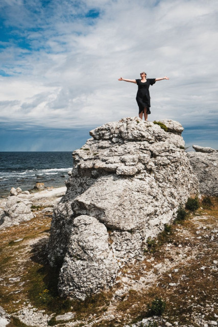 A woman in a black dress stands atop a large weathered limestone rock formation on the Gotland coast, arms outstretched wide with her face tilted upward toward the sky. Her dark hair and dress catch the wind, giving a sense of movement and joy. The rock beneath her is pale, textured, and eroded, part of a group of similar formations along the rocky shoreline. The Baltic Sea stretches out behind her under a partly cloudy sky. Smaller rocks and patches of dry grass fill the lower portion of the frame. The image conveys a sense of freedom and elation against a dramatic natural backdrop.