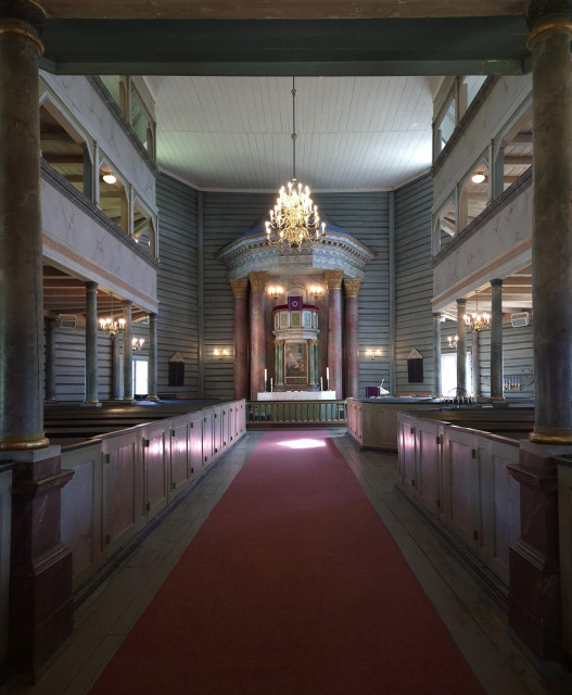 The interior of a typical Norwegian wooden church from the 1830s, featuring doors in front of the pews, a two-storey gallery and a pulpit-altar (a characteristic Norwegian feature from the 19th century). The wooden walls and columns are painted teal, whilst the panelling and the walls of the two galleries are painted in a pink marble-effect finish. The columns in the nave and around the pulpit altar are also painted in a marble-effect finish (those around the altar in pink). A pale pink carpet runs from the entrance to the altar. A brass-coloured chandelier hangs from the ceiling. The church is one of the largest in the region and has a total of 700 seats … just to put that into perspective when compared with churches in other countries.