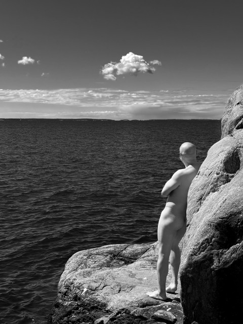 Black and white photo of bald naked man from behind, leaning on a cliff looking out at the sea
