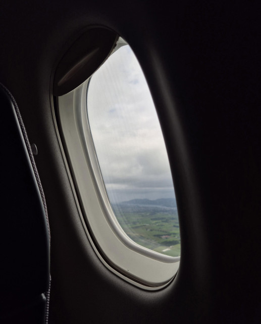 Looking out of an airplane window onto a green landscape and a huge sky covered in white and grey clouds. On the horizon one can see a mountain range and a fjord between the mountain and the flat landscape