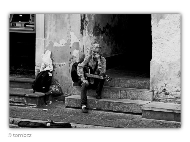 A black-and-white street photograph shows an elderly man sitting on the stone steps of an old building entrance. An acoustic guitar rests across his lap, but he is not playing. He leans slightly forward, one hand covering his mouth while his eyes are cast downward. His deeply lined face, tired eyes, and thoughtful posture suggest a quiet moment of reflection. He wears a checked shirt, dark trousers, and well-worn shoes.
Beside him are a backpack and a white plastic bag resting against a weathered wall with peeling plaster. Behind him, the dark entrance to the building contrasts with the bright, cracked facade. On the pavement in front of the steps lies an open guitar case for donations. Inside it sits a small teddy bear, adding an unexpected touch of warmth to the scene. What draws the eye most is not the guitar, but the man's gesture, his lowered gaze, and his body language. Together, they tell a story of silence, waiting, and the quiet struggle of earning a living one day at a time.
