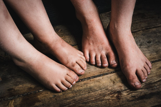 A close-up of several bare children’s feet standing together on a worn wooden floor, dirt visible on the soles and toes from a day spent playing outdoors. The feet are arranged in a loose cluster, overlapping slightly, with visible texture in both the skin and the aged wooden planks beneath. The lighting is soft and natural, with warm skin tones contrasting against the darker wood. The image captures an unposed, everyday moment of summer childhood.