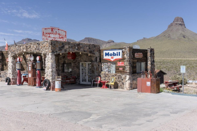 A color landscape photo of a small gas station made of rock. Three old style gas pumps are in front on the left. A very faded sign over the station's pump awning reads, "Cool Springs, Cabins, Tasty Food." The front doors are white. Red and yellow '50s style metal outdoor chairs are seen below a sign on the right side wall that reads, "Mobil." An oil pump island is seen on the far right. The landscape behind the station is desert and an interesting, likely volcanic plug geologic feature is seen on the far right. The sky is bright blue with a few wispy clouds on the far left.