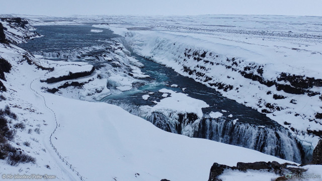 Four colour photos of parts of a frozen waterfall under a snow-laden sky. In the upper left of this shot a wide blue river is making its way towards the viewpoint. Near the centre right it drops over a clutter of eroded boulders and steps, many of which are covered in snow and ice. The canyon wall next to this upper falls is caked with ice. The water course then turns ninety degrees to another drop into a deep canyon, just in front of the viewer. The foreground is a snow field with a large cornice overhanging the canyon, although this isn't obvious from this angle. A line of fencing is running down to a natural rock platform next to the top falls. Compacted ice shows where people have walked down.