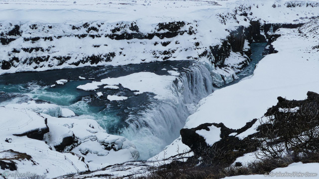 A view from height of the cascade dropping into the gorge. The canyon walls are mostly obscured by ice and snow but dark rock can be seen beneath. The blue water is dropping over a crest which is diagonal to the viewer, a clump of snowy ice is in the centre, splitting the flow. The falling water is white with foam. Opposite the drop, the outline of a cornice is visible, almost completely covered in snow. The foreground is more snowfield with some bare scrub vegetation.