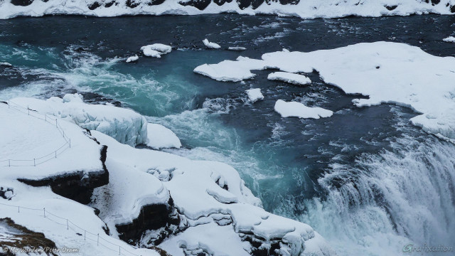 A close-up of the crest of the drop into the canyon. The swirling blue water is white with foam as it passes the natural rock platform which is closed to visitors. Zooming in on the right to 100% of the photo shows footprints all around the edge where visitors have ignored the safety warnings. Icicles and clumps of heavy turquoise ice have formed along the edge of the platform and the snow shows distinct layers.