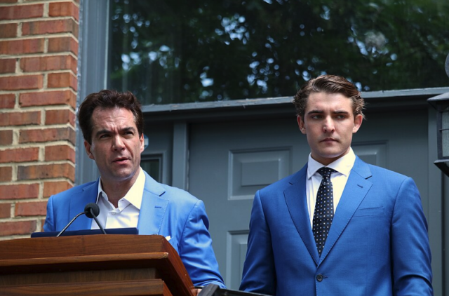 A photo of Burkman (left) and Wohl) outside his Arlington, Va. in a 2020 press conference where they made up sexual assault allegations against public figures. Burkman is and standing at a podium in a blue suit. Wohl, also in a blue suit and tie, stands to the right of Burkman, looking down.