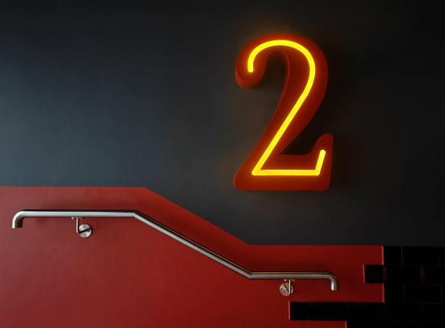 A photo of a glowing neon sign of the number "2" mounted on a dark green wall above a red staircase with metal handrails.