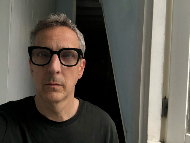 Grey haired man wearing a dark t-shirt and donning thick black eyeglasses is seen near the left side of the frame looking at the camera.
