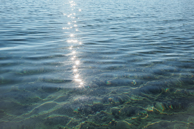 A streak of bright white sun star reflections on the turquoise blue lightly rippled surface of a lake. In the foreground, swirls of sand lit with reflections of the surface under the water. 