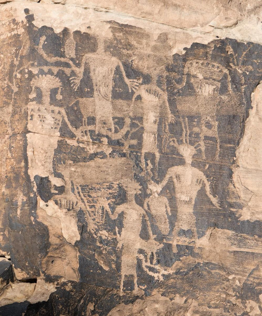 A color portrait photo of a rock imagery panel. The images are carved into a vertical beige colored sandstone rock face that has a very dark desert varnish patina, so the images are light colored and the background is dark. They are a group of overlapping anthropomorphs, each with strange heads or headdresses that have elements that look like horns, feathers, and earrings. (Note: I only describe the images in terms that my modern eye sees them for the benefit of those with limited or no vision. Generally, it is not recommended in Visit With Respect guidelines, established by local Natives, to imbue any meaning upon ancient rock images as we are not of the culture that made them.)