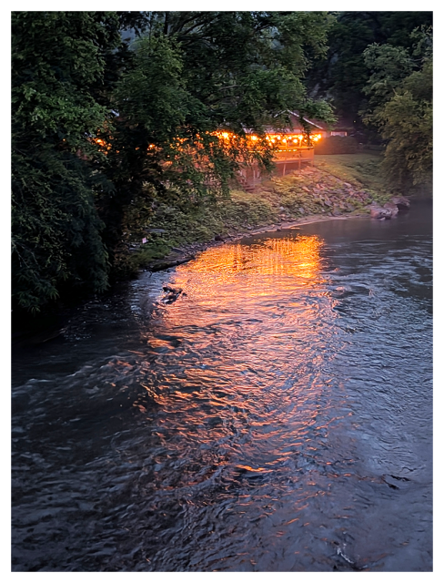 dusk. a river flows beside a wooded bank at dusk. warm yellow lights from a house reflect on the rippling water. trees surround the structure, partially obscuring it.
