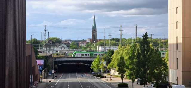 Vordergrund und Perspektive: Der Blickwinkel ist erhöht und schaut mittig auf eine mehrspurige, asphaltierte Straße hinab. Flankiert wird das Bild links von einer dunklen, geriffelten Gebäudefassade und rechts von einem modernen, hellbeigen Bürogebäude.

Mittelgrund: Die Straße führt geradeaus in eine dunkle Unterführung unter einer Bahnbrücke. Auf der Brücke steht oder fährt ein grün-weißer Regionalzug (Eurobahn-Design). Rechts der Straße wachsen dichte, grüne Bäume.

Hintergrund: Hinter den Bahnanlagen erstreckt sich eine grüne Kulisse aus Bäumen und vereinzelten Häusern. Markantestes Merkmal ist ein hoher, historischer Backsteinkirchturm mit einer spitzen, grünlichen Turmhaube, der zentral in den Himmel ragt. Zahlreiche Oberleitungsmasten für die Bahnlinien ziehen sich durch das Bild.

Himmel und Licht: Der Himmel ist bewölkt mit hellen Lücken, das Licht wirkt wie an einem leicht wechselhaften Sommertag.