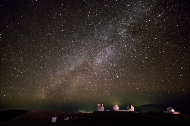 The night sky as seen from the summit area of Maunakea. The Milky Way stretches across the sky above the world-class observatories located there. At the center of the scene are the enclosures for the Subaru Telescope and the Keck I and II Telescopes. To the right, you can see the NASA Infrared Telescope Facility (IRTF), while the James Clerk Maxwell Telescope (JCMT) and the Submillimeter Array (SMA) are in the lower left. Credit text: Subaru Telescope.