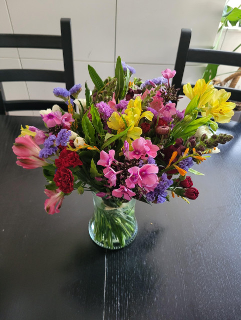 An incredible vase of colourful meadow flowers completely bursting with life and an amazing smell. They're sat on a black wooden table and you can't help but smile looking at or imagining them.