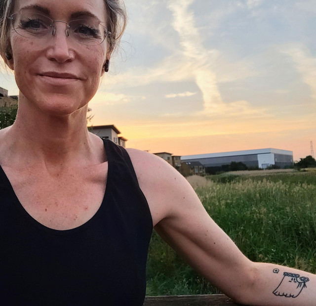 Me standing in front of some marshes on my night walk, with a stunning sunset behind me, it's orange and pink and blue and I look very happy about it.