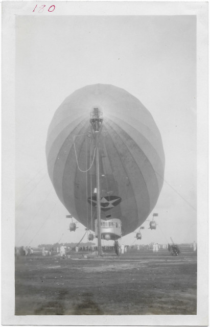 A monochrome, head-on view of the giant air-ship Los Angeles, tethered to a mast just above the ground. In the field below, you can see dozens of tiny people standing, looking.