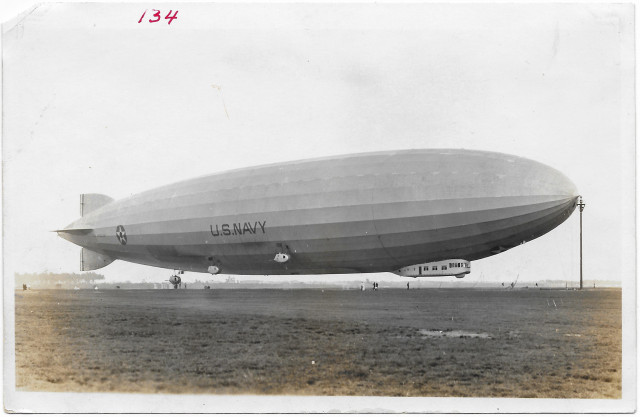 A monochrome, side-view of the US Navy dirigible Los Angleles. Still tethered to a mast, floating maybe15-20 feet off the ground. Less than a dozen people are nearby.