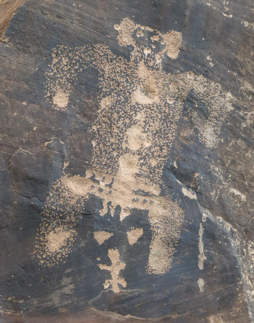 A color portrait photo of rock imagery pecked into a canyon wall. The canyon wall is mostly covered in dark patina but someone hundreds of years ago literally pecked the image of (perhaps) a person with their arms extended and pointing downward and their legs spread wide and similarly pointing downward in a ninety degree fashion. The pecking created a salt and pepper look in the figure's body. The head has eyes and a mouth and prominent ears sticking out.  (Note: I only describe the image as they are regarded in modern culture for the benefit of those with limited or no vision. Generally, it is not recommended in Visit With Respect guidelines, established by local Natives, to imbue any meaning upon ancient rock images as we are not of the culture that made them.)