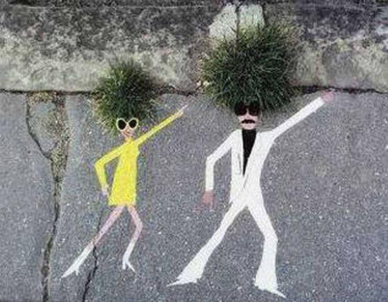 Twee pollen onkruid tussen de stoeptegels vormen de haardossen van twee dansende figuurtjes die met krijt eronder zijn getekend.
