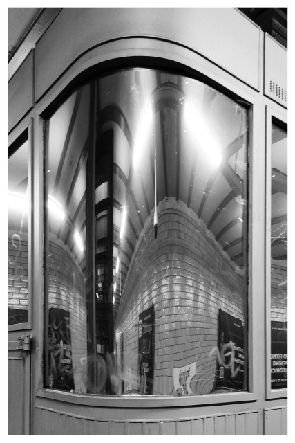 Schwarz-Weiß-Foto im Hochformat mit weißem Rand. Zu sehen ist die gebogene spiegelnde Scheibe eines Aufsichtshäuschens auf einem U-Bahnsteig. In der gebogenen Scheibe spiegeln sich die gekachelten Wände ebenfalls gebogen.