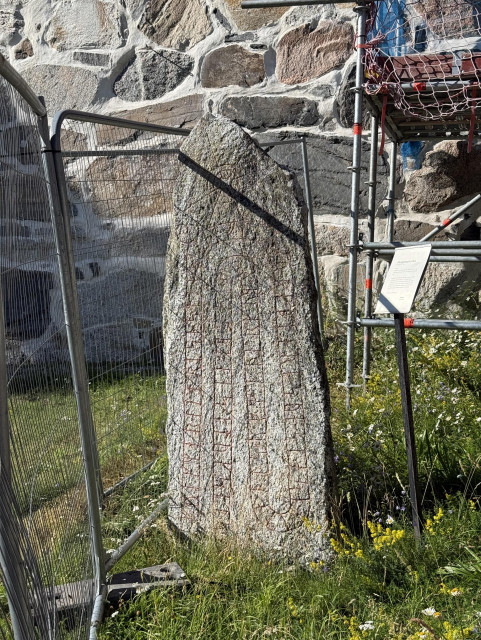 Photo of a runestone in front of a stone wall surrounded by fences and scaffolding. 