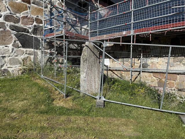 Photo of a runestone behind a fence in front of scaffolding next to stone walls. 