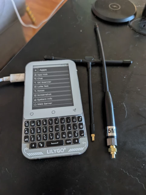 This image displays a small handheld communication device, the tdeck max, and its accessories on a dark surface. The device is centered, light gray with a white screen and a black QWERTY keyboard. A white USB cable connects to its left side. The screen displays a header, Apps, followed by a list of applications, App Hub, Chat, I2C Scanner, LoRa Test, Notes, Screenshot, System Info, and Web Server. Below the screen are three circular buttons, then a physical QWERTY keyboard with black keys. At the bottom of the device, LILYGO  is printed. To the right of the device are two black antennas,a T antenna and a long one. The longer antenna has a silver band with the numbers 915M near its base.