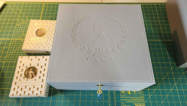 A pale blue jewelry box with a golden key. It has the silhouette of a head surrounded by a wreath with a bow embossed on it. Beside it are two small boxes for the Trip to Bath and Season in London expansions.