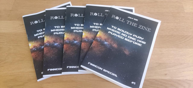 Five copies of the same zine fanned out on a table. The zine is Roll the Zine's July 2026 issue, a Finncon special: To boldly play where no one has played before.