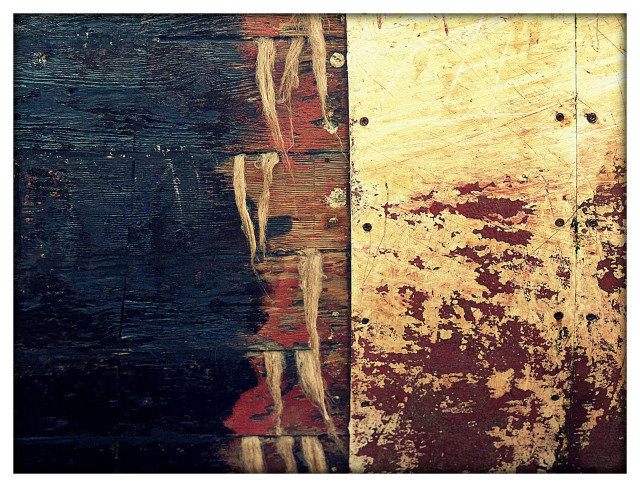 A colour photograph showing a small section of trawler hull under repair. A vertical band of prussian blue dominates the left f the frame and a combination of broken red and cream steel hull dominates the right. The centre of the frame shows wooden hull slats with filling material hanging down from the seams.
