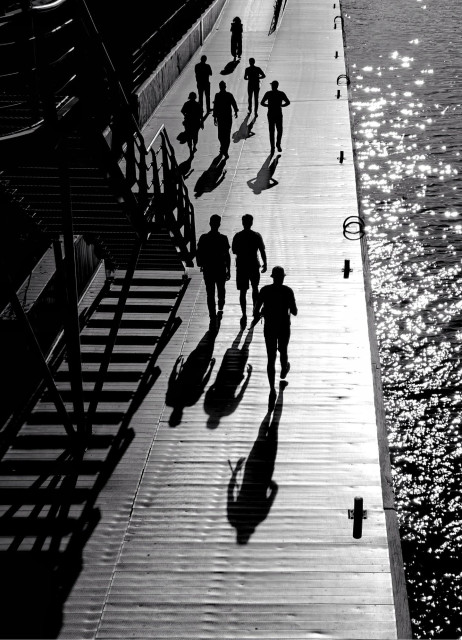 A black-and-white photo looking down on several  people walking on a waterfront promenade, casting long shadows over the bright light reflected by the water and pavement.