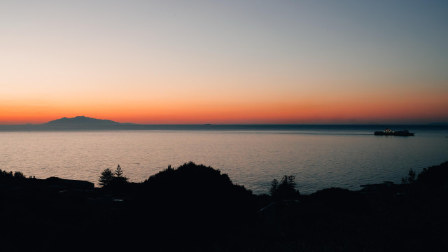 A sunrise over calm mediterranean waters, with silhouettes of hills and dark foliage in the foreground. A distant island (Elba) is visible on the left, while on the right a ferry boat can be seen sailing across the sea towards the port of Bastia, Corsica. The sky exhibits a gradient of warm colors from orange to purple.