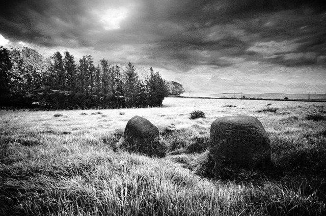 Atmospheric black and white photo of two small standing stones in a grassy field, with trees in the middle distance, under dark clouds.