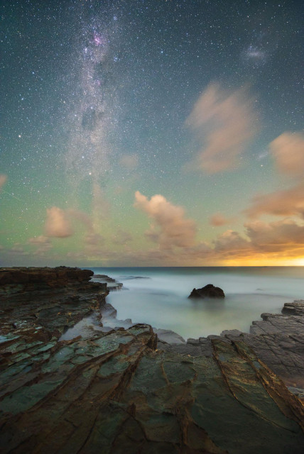 night seascape with rock platform, lone submerged rock and milky way above it all. Some smoother clouds and some light pollution looking like sunrise or sunset on the right hand side.