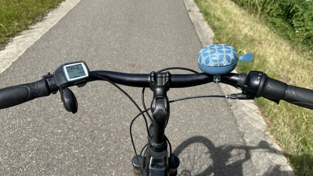 View of a bicycle handlebar with a digital speedometer, a blue patterned bell, and a small blue bike charm, on a paved path with grassy sides.