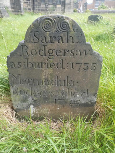 An upright gravestone in a grassy graveyard, the stone is in very good condition for its age. There is an intricate circular pattern at the top, then the wording Sarah Rodgers was buried 1735 Marmaduke Rodgers died 1855.